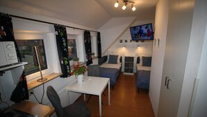 A compact, well-lit room with a kitchenette, a dining area, and a TV mounted on the wall.
