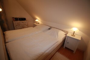 Un letto singolo con lenzuola bianche in una stanza con soffitto spiovente. Due comodini con lampade sono posti ai lati del letto, uno per parte. Un settimino in legno è appoggiato alla parete a sinistra del letto.