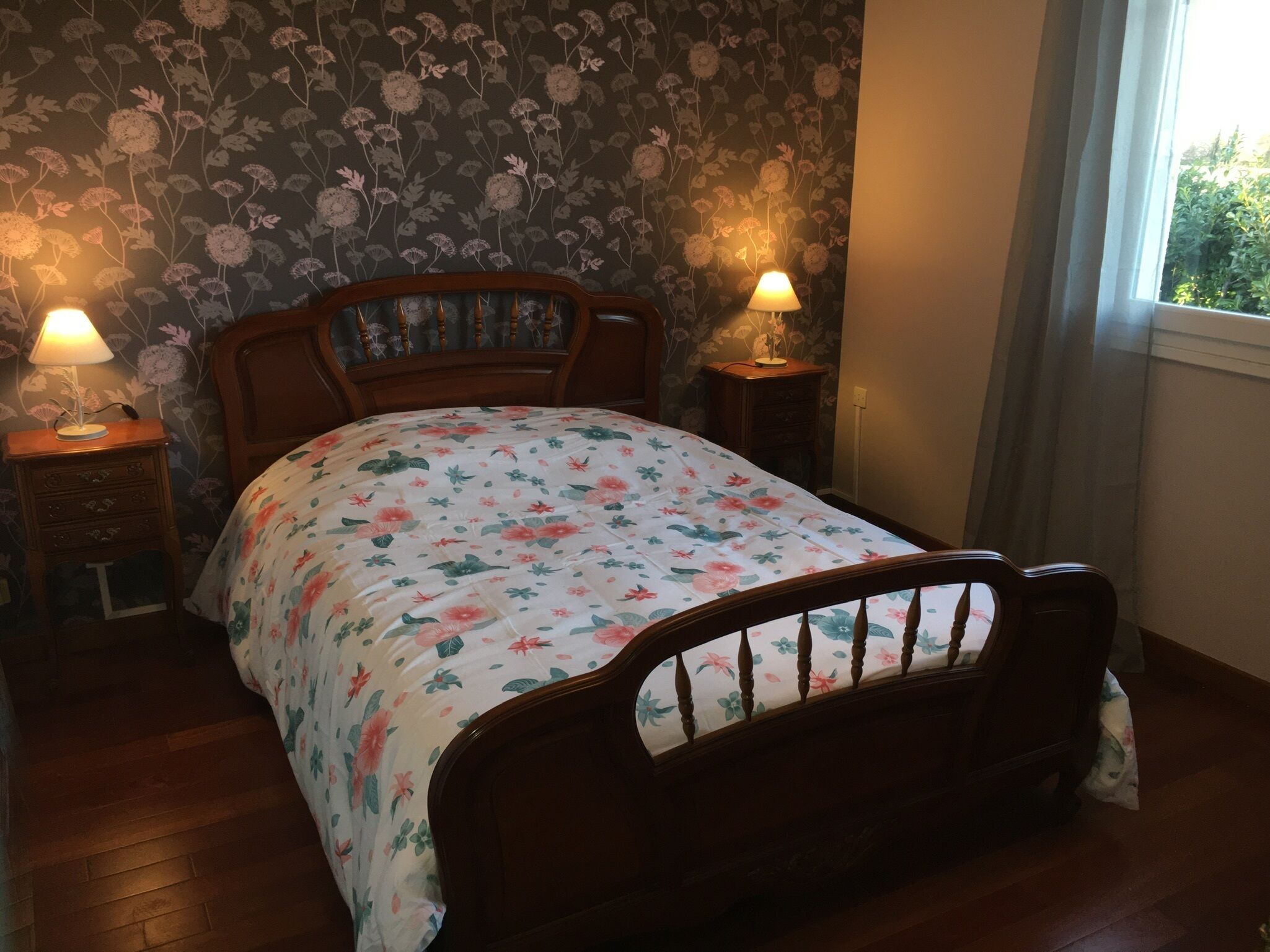 Een slaapkamer met een bloemenbedekking, houten hoofdeinde, twee bedlampjes en een raam met gordijnen.