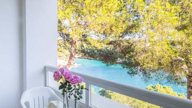 Ein Balkon mit Tisch, einer Vase mit rosa Blumen und Blick auf ein türkisblaues Meer und grüne Bäume.