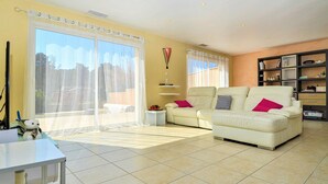 A spacious living room with a large sliding glass door leading to a balcony, a white sofa with cushions, and a bookshelf filled with books and decorative items. - Amazing home in Mornas (Mornas)