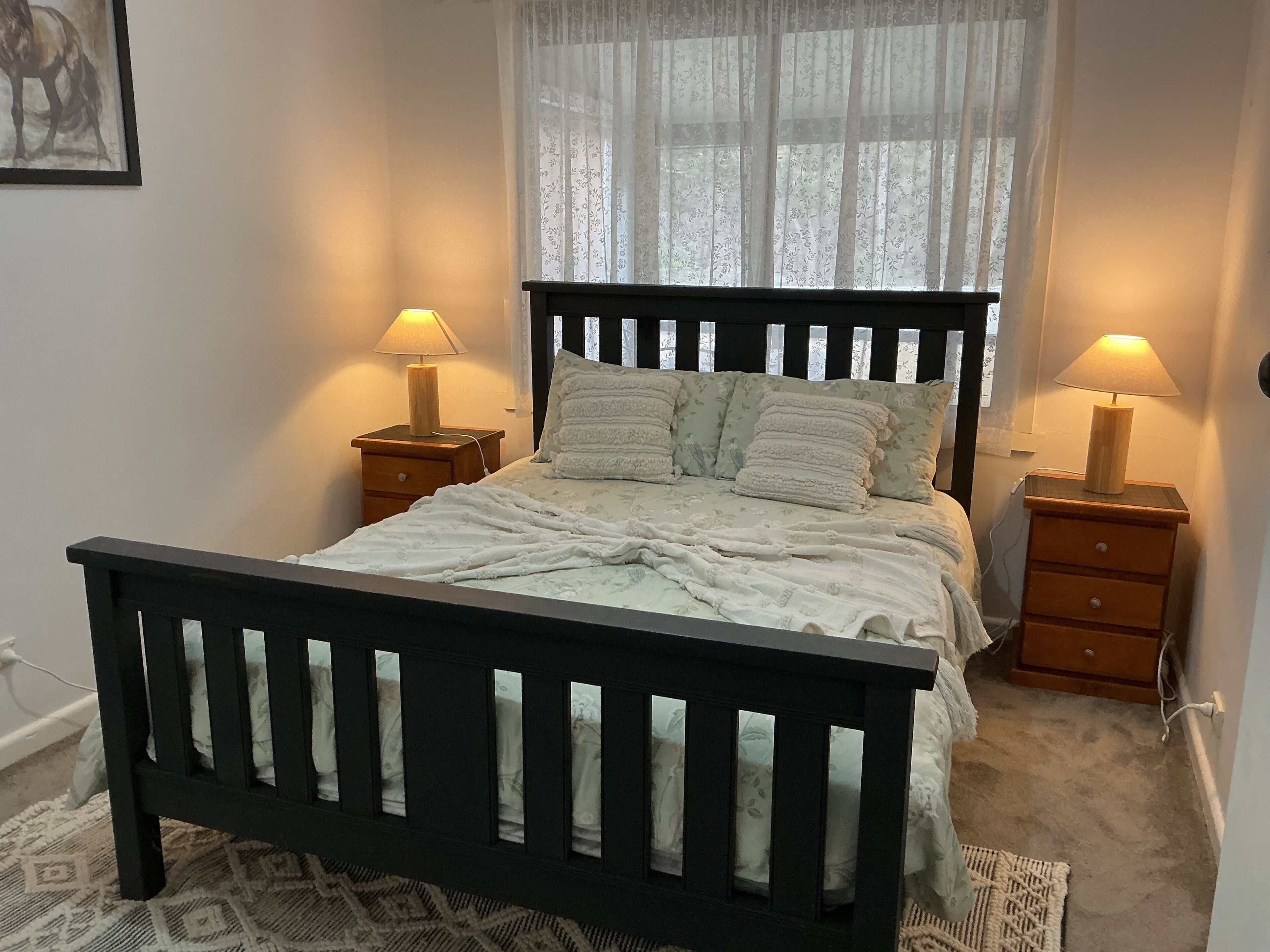 A bedroom with a wooden bed, two bedside tables with lamps, a window with sheer curtains, and a patterned rug.