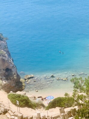 A coastal beach with clear blue water, rocky shoreline, and a few people in the distance.