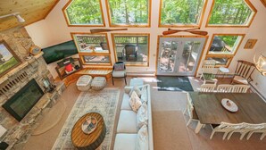 A spacious living area with a stone fireplace, a large sofa, a dining table, and multiple windows offering views of a forest. - Dockside Dog Friendly Retreat w/ Views & Arcades (Swanton)