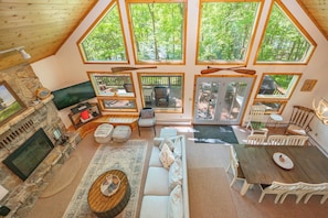 A spacious living area with a stone fireplace, a large sofa, a dining table, and multiple windows offering views of a forest.