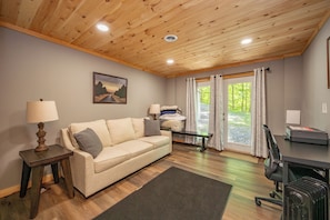 A room with a wooden ceiling, a sofa, a desk, a lamp, a chair, and a window with curtains.