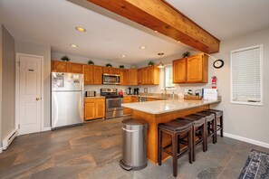 A modern kitchen with wooden cabinets, stainless steel appliances, a central island with stools, and a stone tile floor.