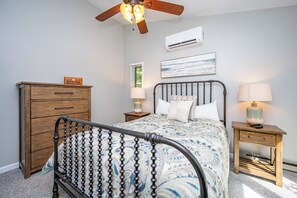 A bedroom with a bed, wooden dresser, nightstands, a ceiling fan, an air conditioner, and a window with blinds.