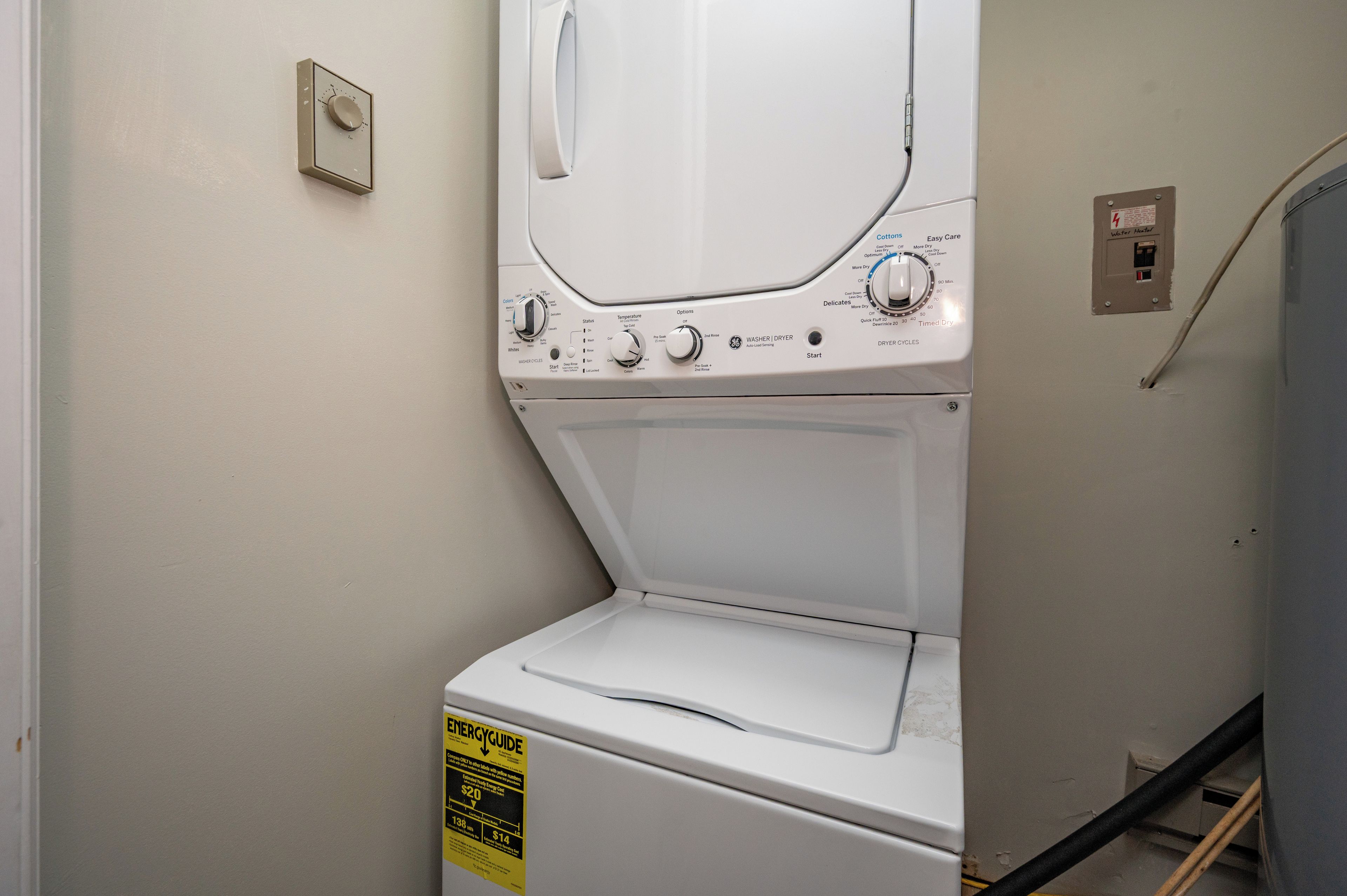 A white front-loading washing machine with a control panel, a power outlet, and a water heater switch on the wall.