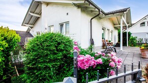 Une maison blanche avec un toit en pente, une véranda et un jardin parsemé de fleurs roses.