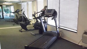 Ein Fitnessraum mit Laufbändern und Ergometer, großen Fenstern mit Jalousien und einem Teppichboden.