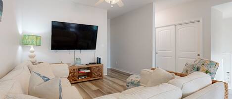 A living room with a large flat-screen TV, a ceiling fan, a wooden entertainment unit, and a wicker chair.