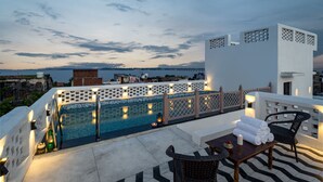 Zona de piscina en la azotea con vista al mar y un muelle distante, que incluye un edificio blanco, una piscina con borde de azulejos y mobiliario exterior.