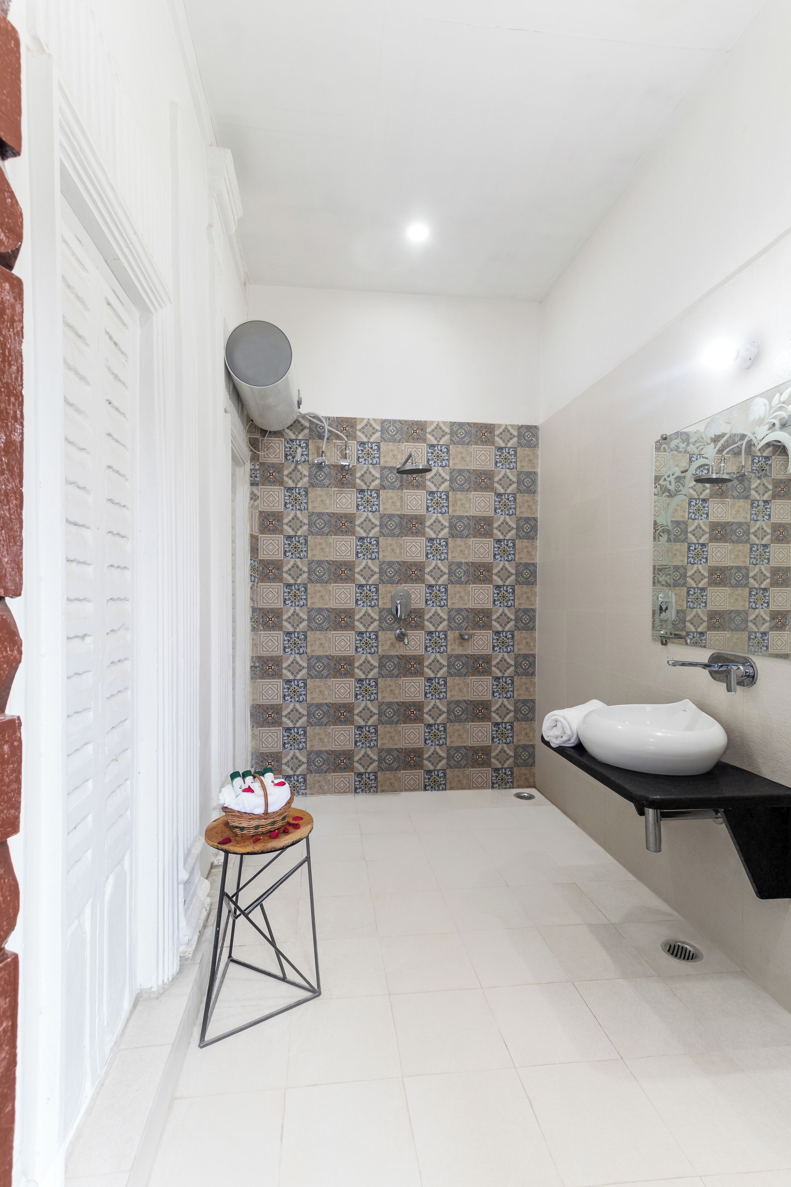 A bathroom with a white tiled floor, a patterned tile shower wall, a white sink with a black countertop, and a small stool with a basket.