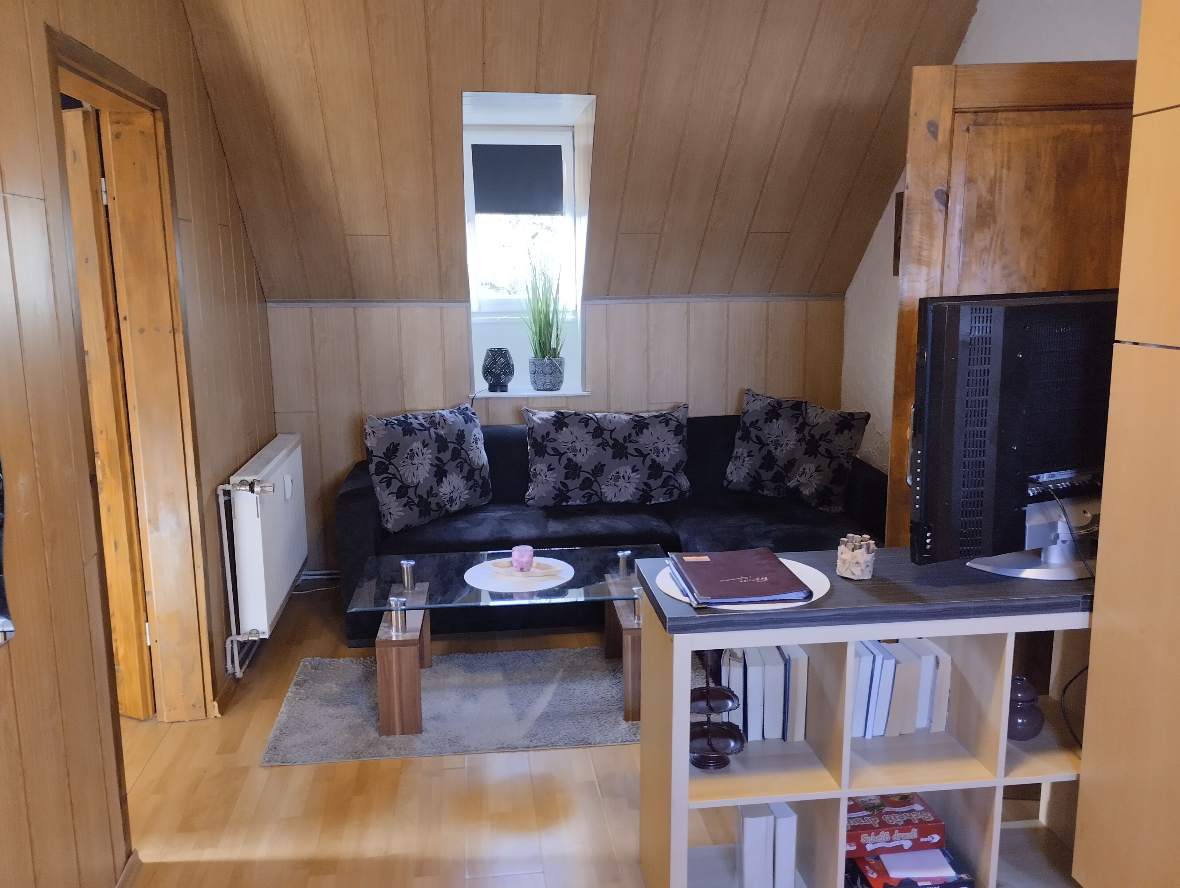A cozy living room with a wooden ceiling, a black sofa with floral cushions, a glass coffee table, and a bookshelf.