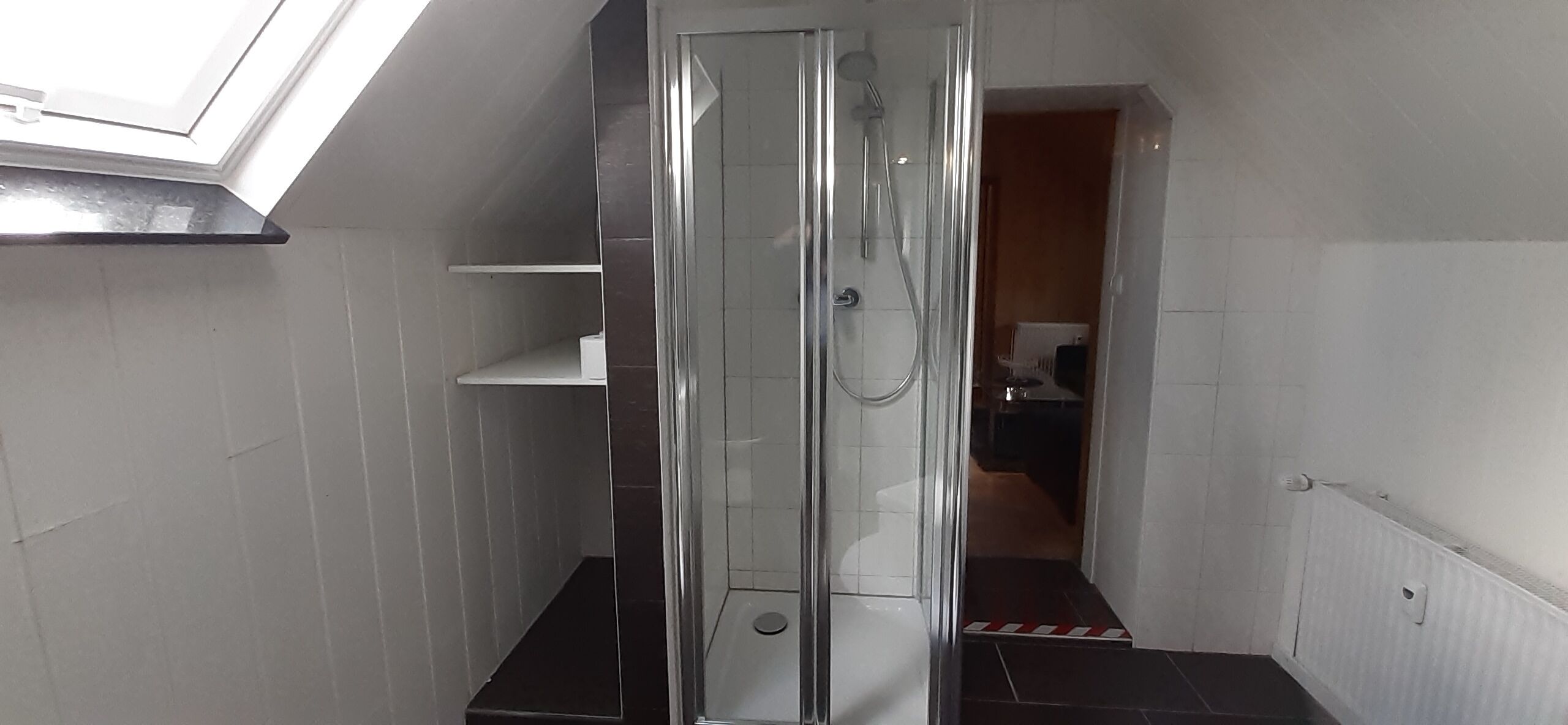 Une petite salle de bain avec une douche à parois de verre, des murs recouverts de carreaux blancs et une petite étagère.
