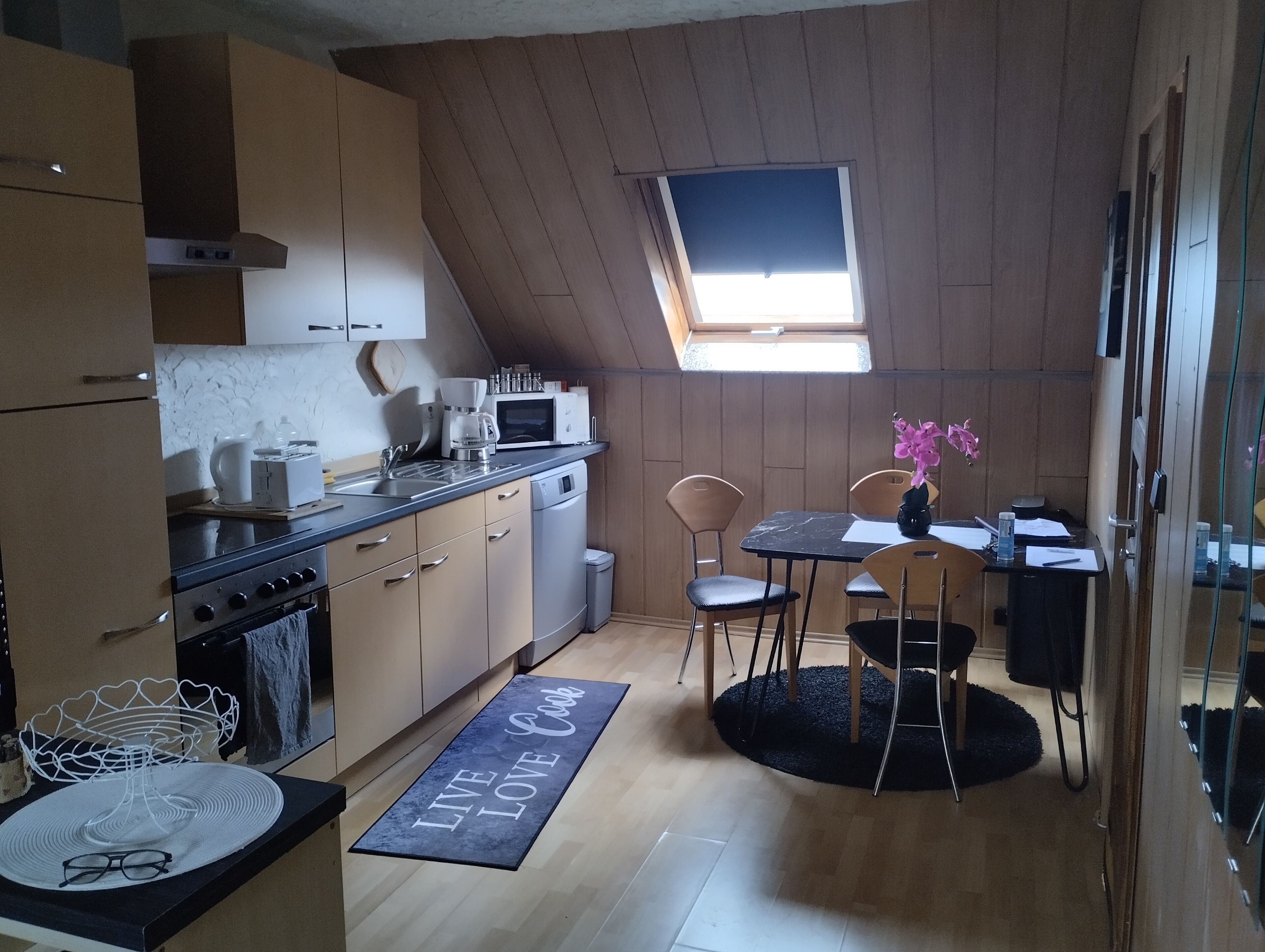 Une cuisine compacte dotée d’un puits de lumière, un coin repas avec un tapis noir et une petite table accompagnée de chaises.