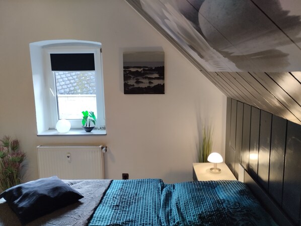 A bedroom with a bed, a window with a view, a radiator, a wall-mounted lamp, and a painting. - Ferienwohnung "Am Vulkanpfad" (Ettringen)