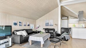 Ein modernes Wohnzimmer mit Flachbildfernseher, einer Couch, einem Couchtisch und einem Essbereich mit Stuhl sowie einer Küche im Hintergrund.