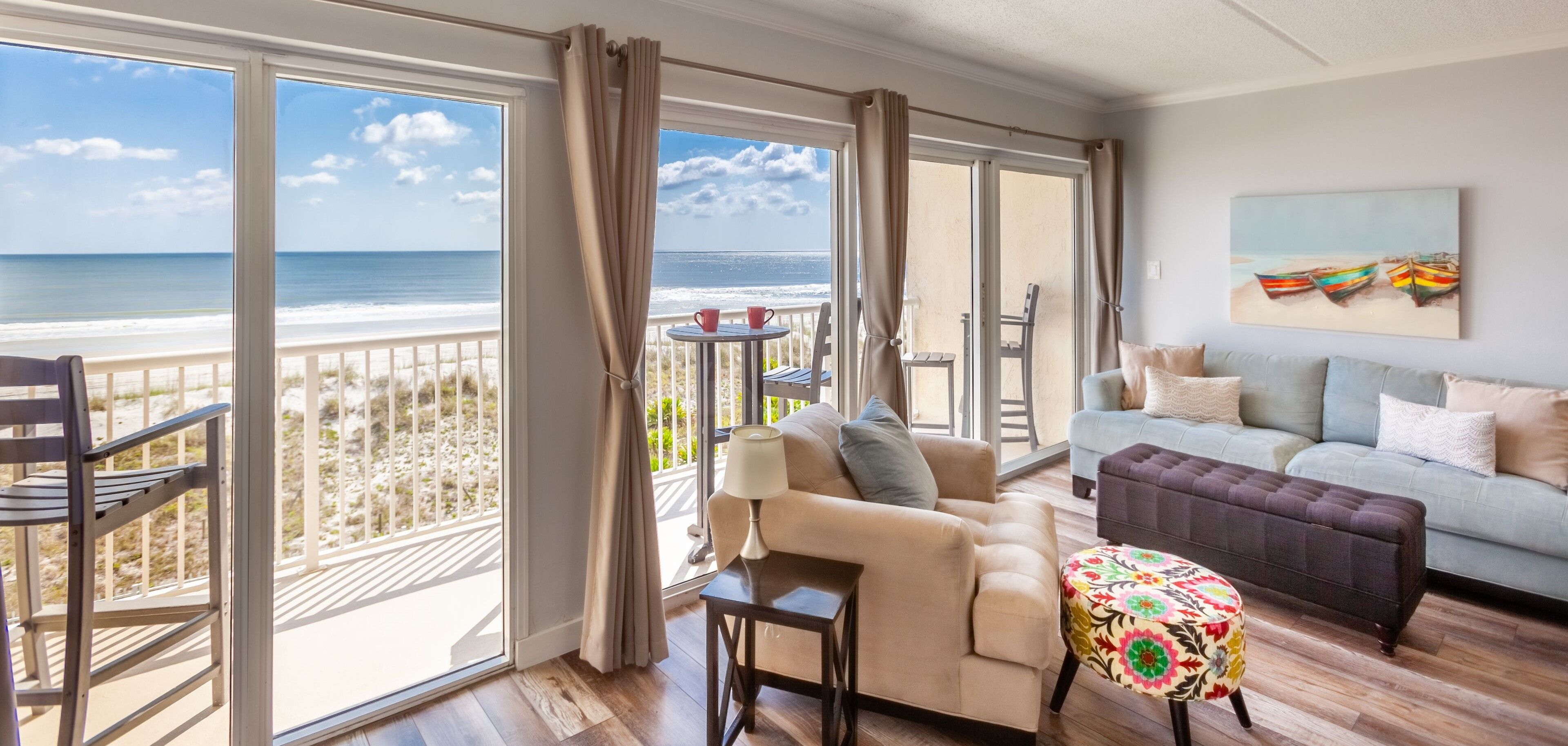A room with a balcony overlooking the beach, a sofa, a small table, and a colorful ottoman.
