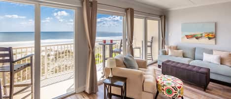 A room with a balcony overlooking the beach, a sofa, a small table, and a colorful ottoman.