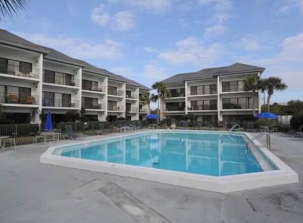 A swimming pool surrounded by a multi-story building with balconies, a clear sky, and palm trees.