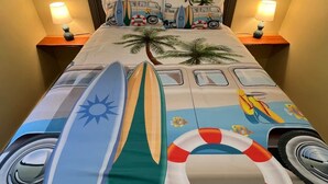 Un lit décoré sur le thème d’une plage tropicale, avec des planches de surf, une bouée de sauvetage et un van orné de palmiers.