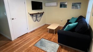 Un espace de vie compact doté d’un plancher en bois, d’un canapé noir avec des coussins turquoise, d’une petite table, d’un hamac, d’un téléviseur et d’un appareil de climatisation.