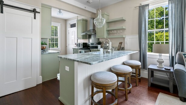 A kitchen with a central island, bar stools, a hanging light fixture, and a window with curtains. - The Jasmine Suite | 3 Bedrooms Steps from Upper King (Charleston)