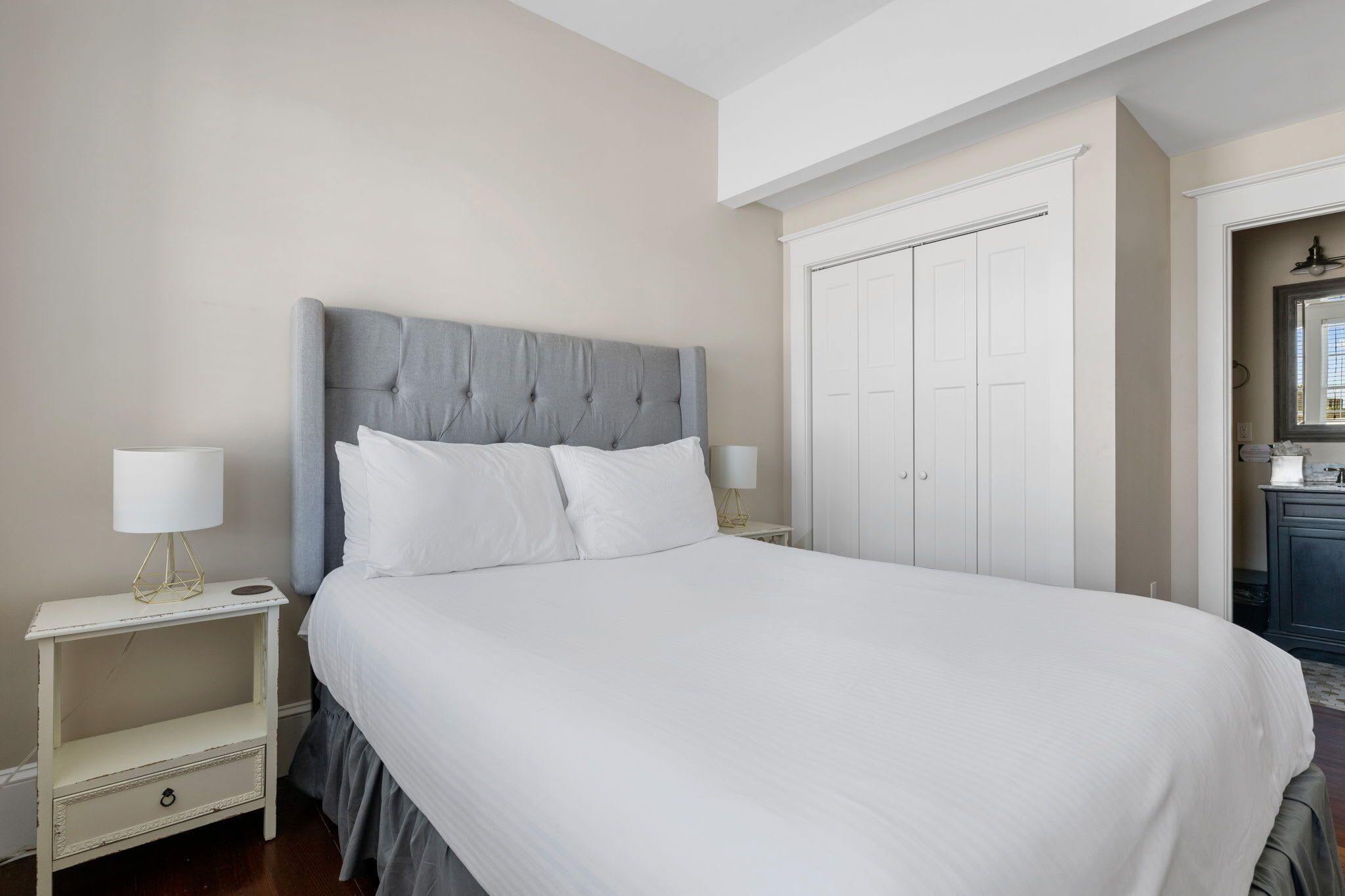 A neatly made bed with a grey headboard, a white nightstand with a lamp, and a wardrobe with a mirror.