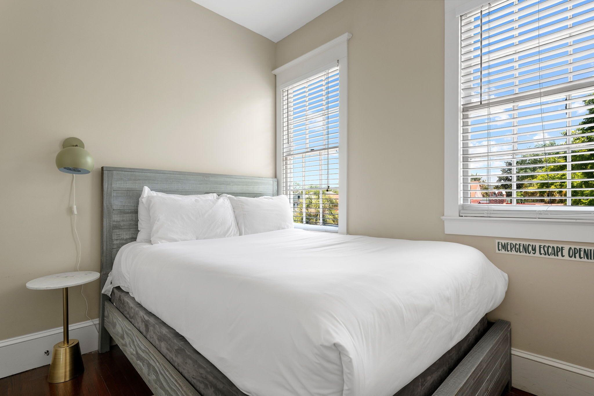 A bedroom with a bed, a bedside table, and a lamp. There is a window with blinds and a sign indicating an emergency escape route.