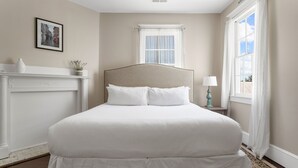 A neatly arranged bedroom with a large bed, a small bedside table, a nightstand, a window with curtains, and a framed picture on the wall. - The Jasmine Suite | 3 Bedrooms Steps from Upper King (Charleston)