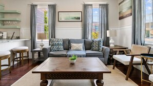 A living room with a sofa, coffee table, and chairs. There is a window with curtains, a painting on the wall, and a plant on the coffee table. - The Jasmine Suite | 3 Bedrooms Steps from Upper King (Charleston)