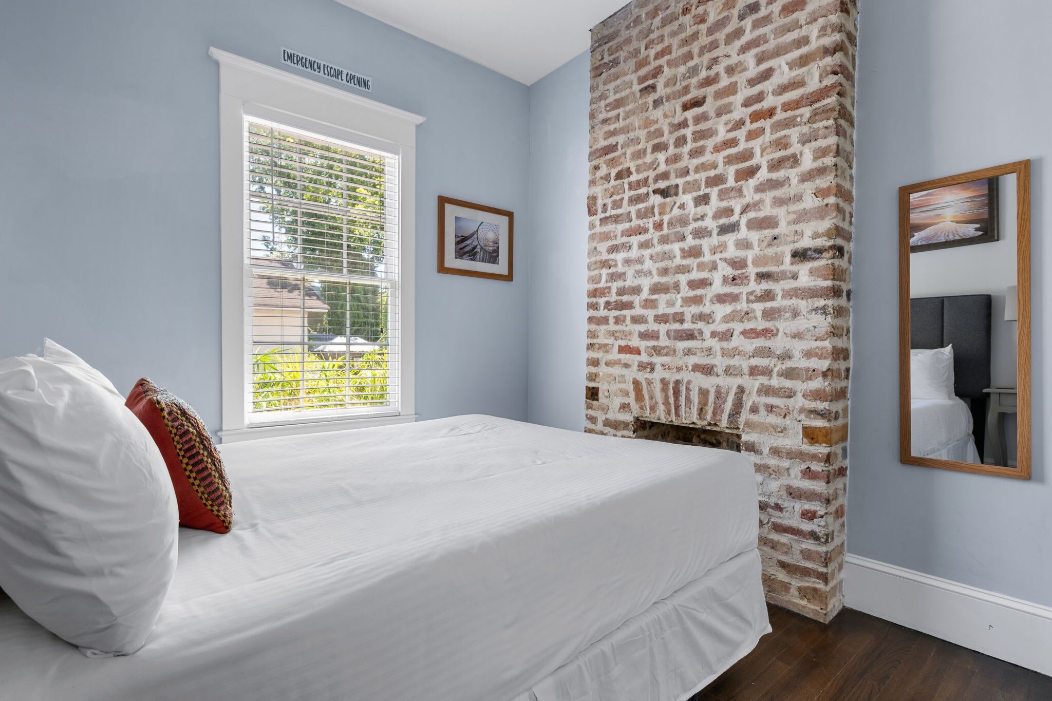 A bedroom with a brick wall, a window with blinds, a bed with white bedding, and a framed picture on the wall.
