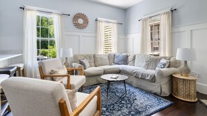 Een woonkamer met een beige hoekbank, een ronde salontafel en een blauw tapijt met patroon.