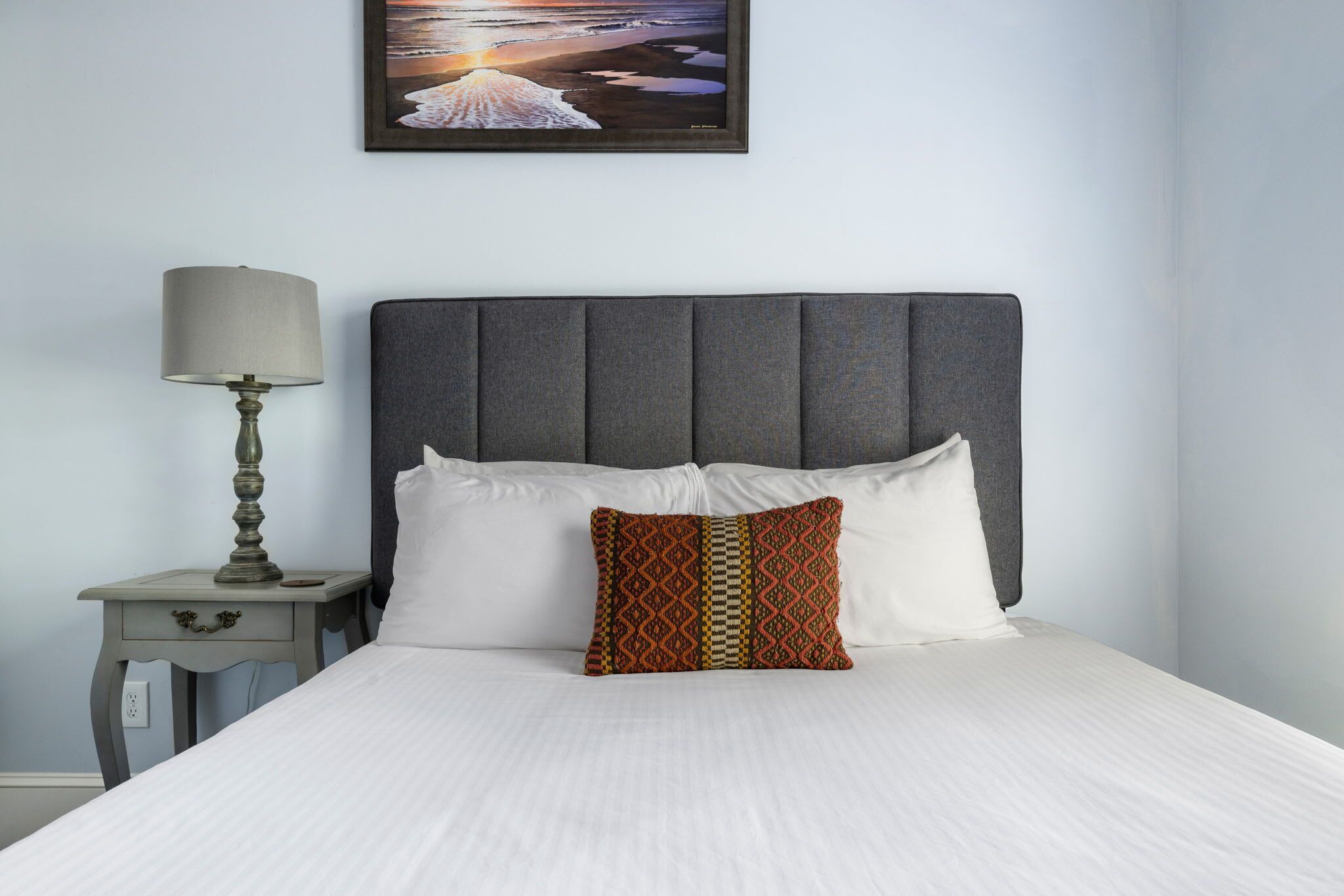 A neatly made bed with a patterned pillow, a bedside lamp, and a framed picture on the wall.