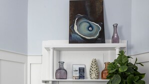 Een witte open haard met daarboven een schilderij, een plank met decoratieve objecten, en een potplant aan de rechterkant.