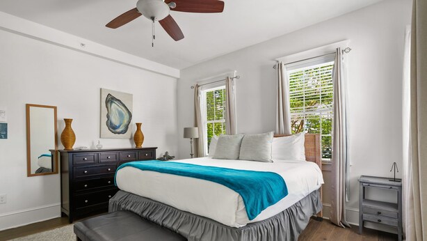 A bedroom with a bed, a dresser, a bench, a ceiling fan, and a window with curtains. - The Oyster Suite | Walk to King + Cannon Green (Charleston)