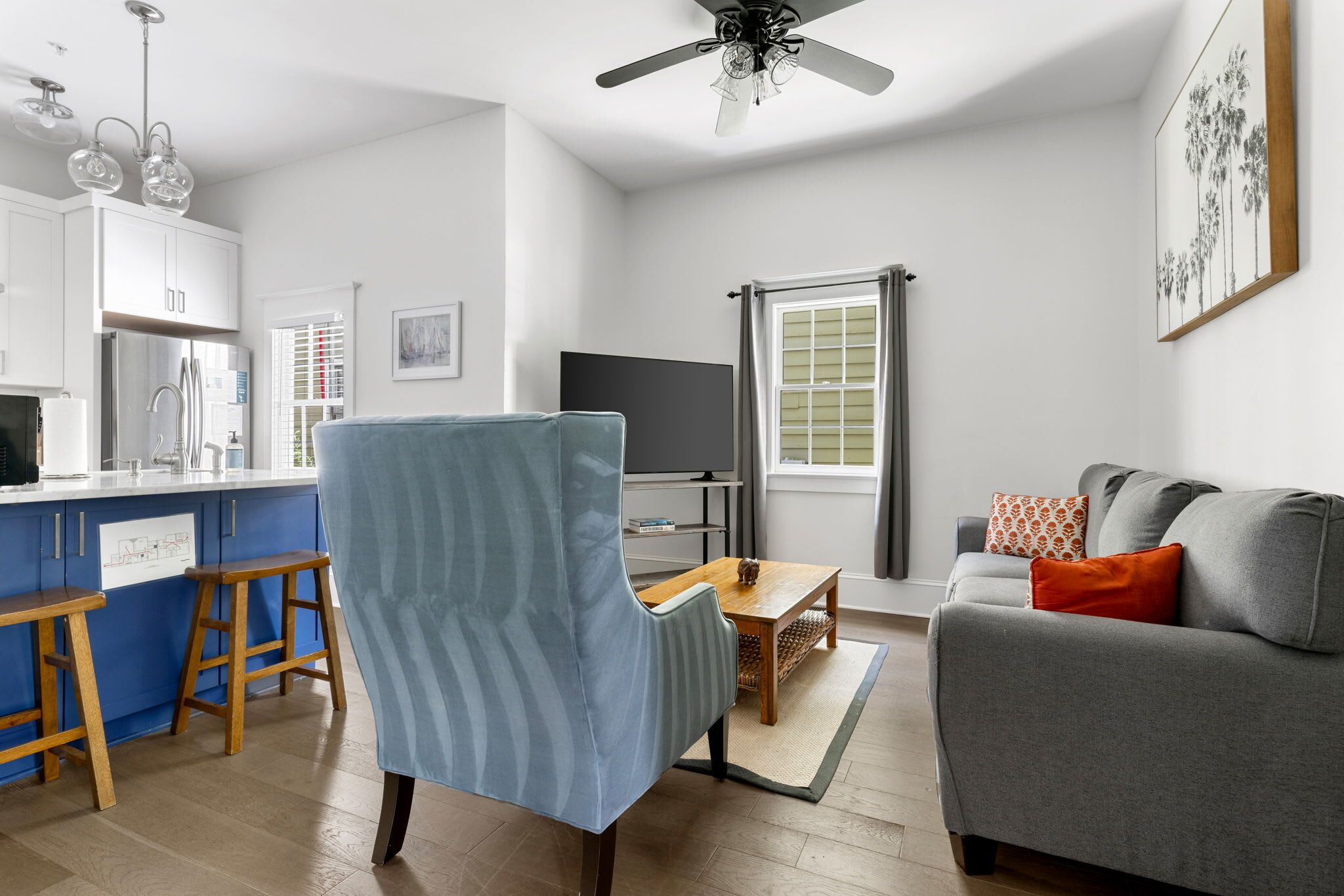 A modern living room with a grey sofa, a blue armchair, and a wooden coffee table. There is a kitchen area with white cabinets and a blue bar stool.