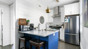 A modern kitchen with white cabinetry, a central island with stools, a stainless steel refrigerator, and a hanging pendant light. - The Hydrangea Suite | 3BR/2BA | Sleeps 6 (Charleston)