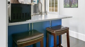 A kitchen with a white countertop, two wooden barstools, a black appliance, a stainless steel sink, and a hanging pendant light. - The Hydrangea Suite | 3BR/2BA | Sleeps 6 (Charleston)