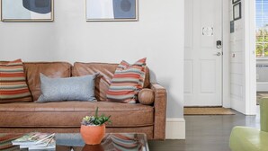 A modern living room with a brown leather sofa, colorful pillows, a glass coffee table with magazines and a plant, and abstract art on the wall. - The Hydrangea Suite | 3BR/2BA | Sleeps 6 (Charleston)