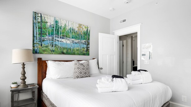 A neatly made bed with white linens and towels, a painting of a forest scene on the wall, and a small bedside table with a lamp. - The Hydrangea Suite | 3BR/2BA | Sleeps 6 (Charleston)