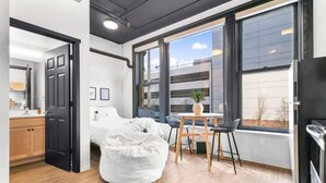 A modern bedroom with a bed, a round bean bag chair, a small table with a plant, and a view of the outside through large windows. - Historic Studio w/ Lux Mattress Walk 2 Everything! (Spokane)