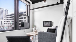 Une chambre d’hôtel moderne dotée d’une grande fenêtre, d’un téléviseur à écran plat, d’un canapé gris, d’une petite table et d’une chaise noire.