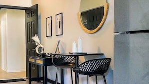 Une cuisine avec un plancher en bois, un réfrigérateur, un miroir rond et deux chaises noires accompagnées d’une table.