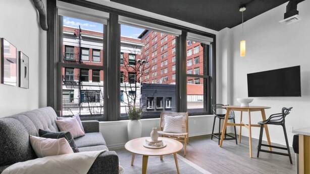 A modern living room with a grey sofa, a round wooden coffee table, and a wicker chair. Large windows offer a view of red brick buildings. - Clean Upscale Hideout Close to Everything + WiFi (Spokane)