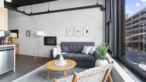 A modern living room with a grey sofa, a wooden coffee table, and a wicker chair. There is a large window with a view of a parking garage. - Clean Upscale Hideout Close to Everything + WiFi (Spokane)