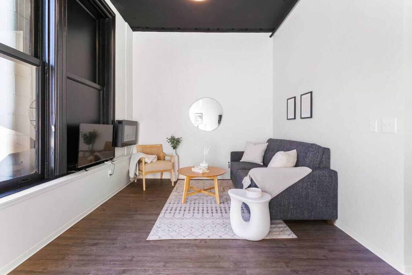 Un salon moderne avec un canapé gris, une chaise en bois, une petite table ronde et une table d’appoint blanche. Une grande fenêtre se trouve à gauche et un miroir est accroché au mur.
