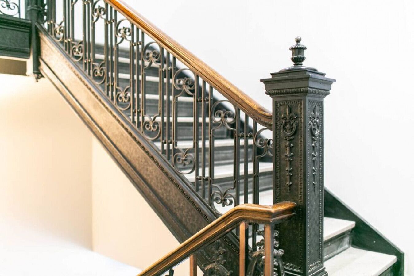 Un escalier orné de rampes en métal et doté d’une main courante en bois, menant à un palier agrémenté d’une colonne décorative.